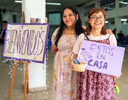Two women warmly greet visitors at a community event in Ciudad de México with signs saying 'Bienvenidos' and 'Estás en Casa'.