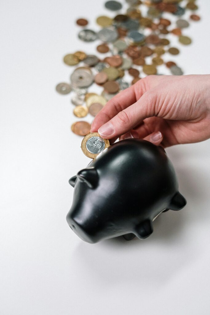 pexels-photo-3943724-3943724 A hand putting a coin into a black piggy bank with scattered coins on a white background.